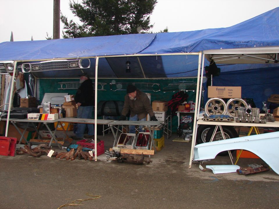 Swap meet booth under blue tarp with chrome wheels and auto parts on tables
