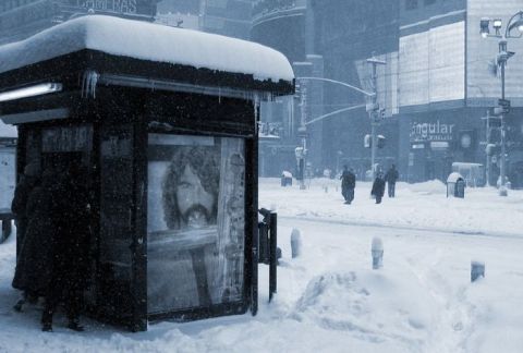 1990 Ford Bronco - A snowstorm blankets a city street, with a bus shelter and pedestrians visible amidst the falling snow.