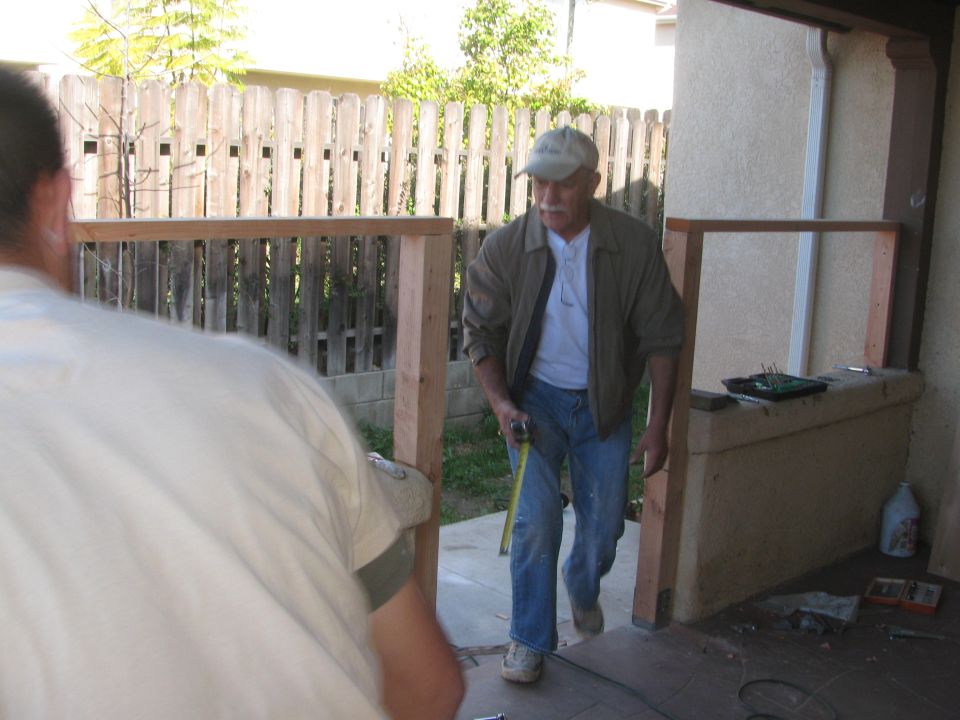 Two men are working on a wooden frame structure outdoors, one is measuring with a tape measure