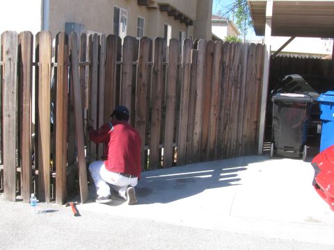 1968 Ford Mustang - A person works on a wooden fence in a driveway, with trash bins and a red car part in the background.