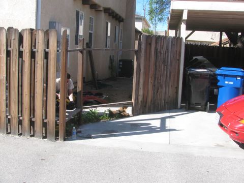 1968 Ford Mustang - A red car is partially visible in the foreground, parked next to trash bins and a wooden fence.