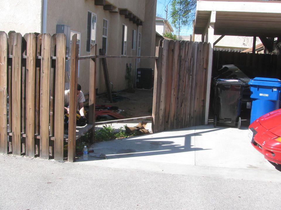 Red car front corner visible, parked next to trash cans and a wooden fence