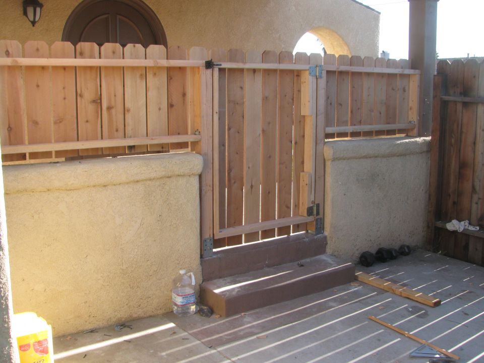 Wooden fence with a gate, steps leading up to it, and a stucco wall. Dumbbells are on the ground