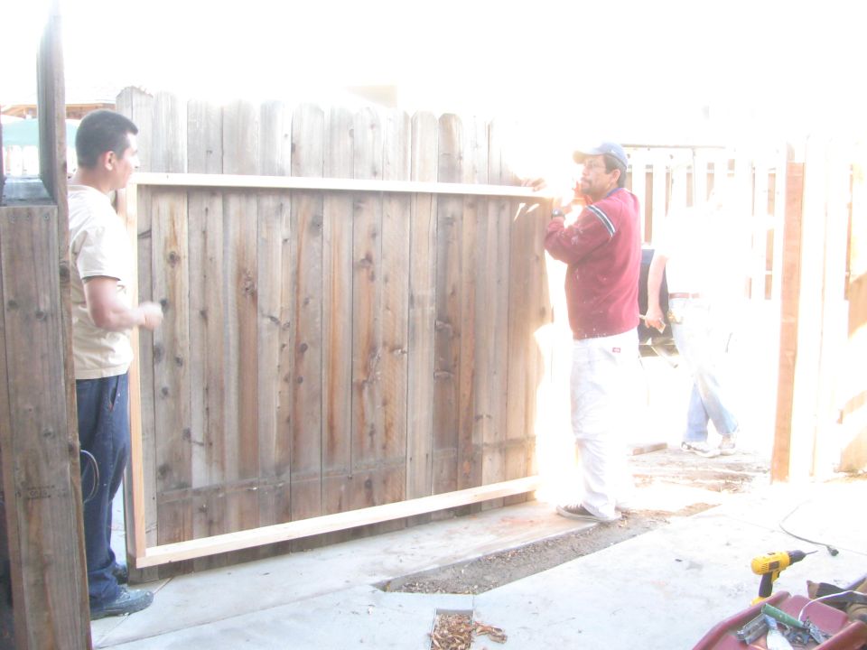 Two men are installing a large wooden fence panel. One man on the left is holding the panel, and the other man on the right is adjusting it