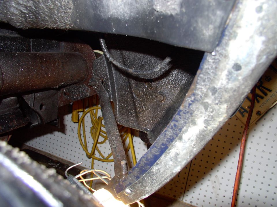 Underside of car showing rusty undercoated frame and wheel well area, blue paint on fender lip, garage pegboard behind