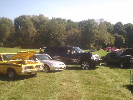 Several cars parked on grass at an outdoor event, with trees in the background. One yellow car has its hood open