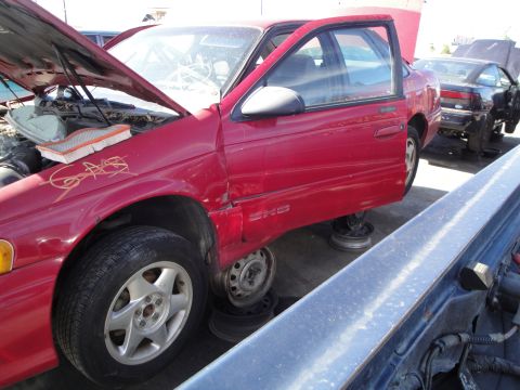 1993 Ford Taurus - A red sedan, possibly a 1993 Ford Taurus SHO, sits in a junkyard with its hood and door open and missing wheels.