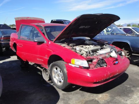 1993 Ford Taurus - A red 1993 Ford Taurus SHO sits in a junkyard with its hood open and missing front wheels.