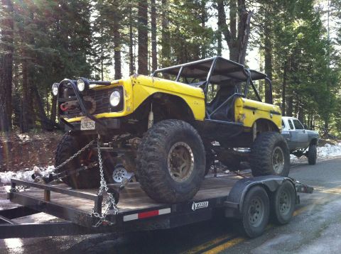 1989 Ford Bronco - Modified yellow off-road vehicle with a roll cage is transported on a trailer, with another vehicle visible on the road behind.