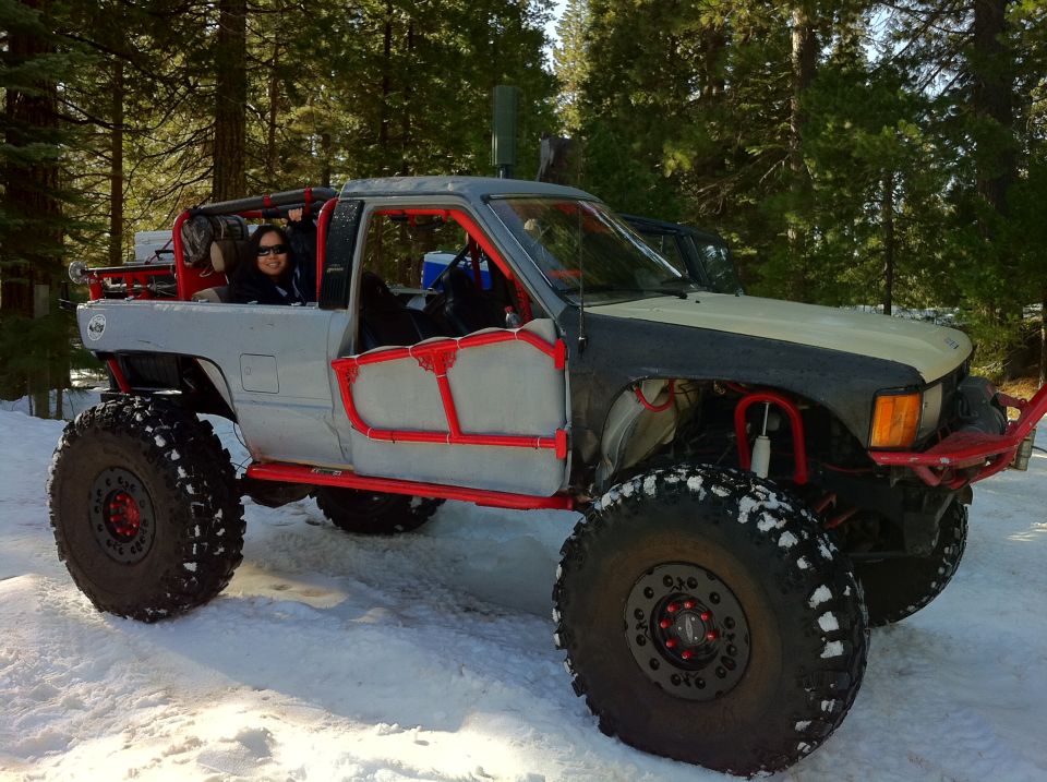 Modified pickup truck with large tires and roll cage in a snowy forest. A person is in the driver's seat