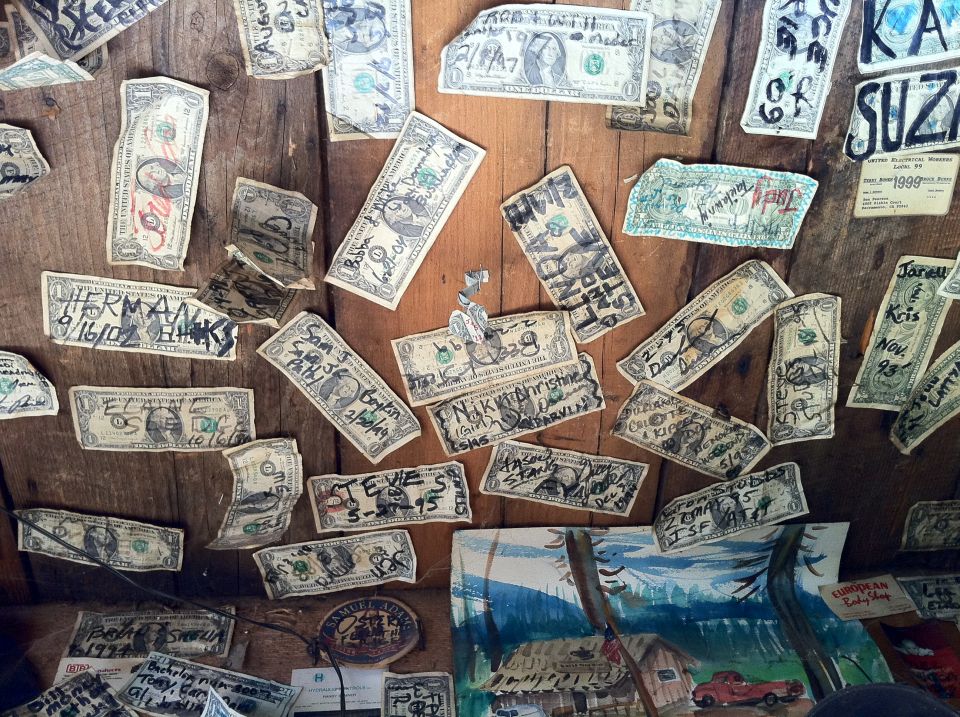 Dollar bills with names and dates written on them, pinned to a wooden wall