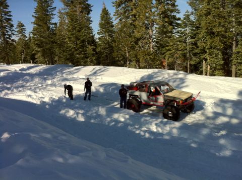 1989 Ford Bronco - A heavily modified off-road truck with oversized tires is stuck in the snow, with several people attempting to pull it free.