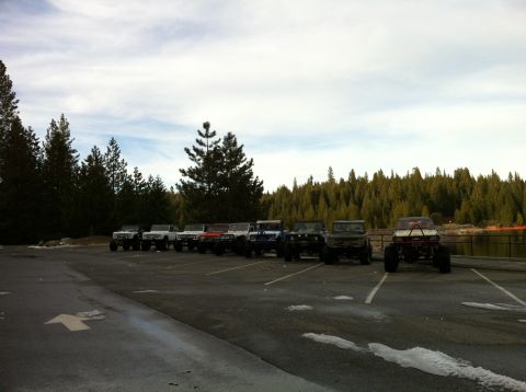 1989 Ford Bronco - A line of off-road vehicles are parked in a lot near a wooded area and a body of water.