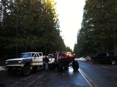 1989 Ford Bronco - A lifted pickup truck and a custom off-road vehicle are parked on a wet road lined with pine trees.