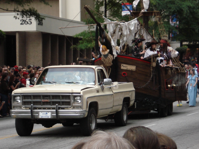 Tan GMC squarebody pickup towing a pirate ship parade float on a city street