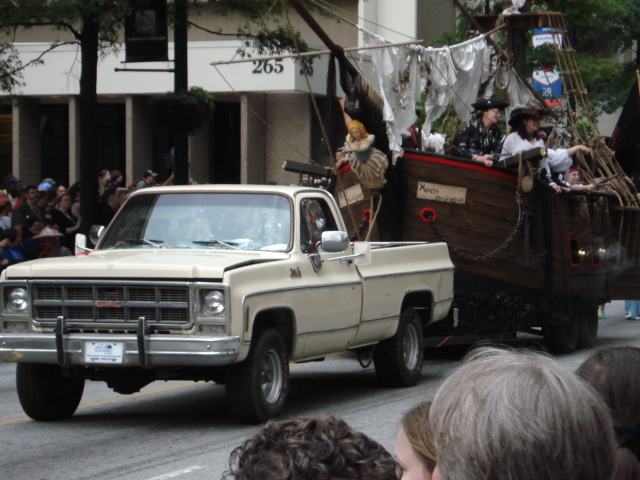 Cream square-body GMC pickup towing a pirate ship parade float past a crowd