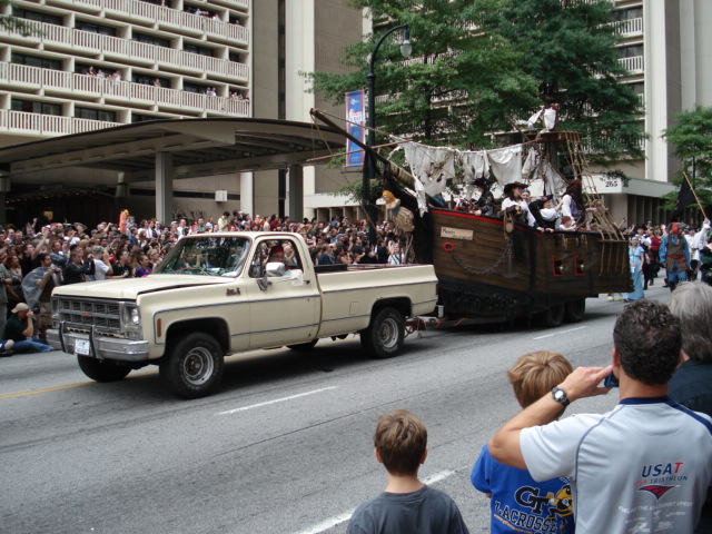 Cream and tan square-body GMC pickup towing a pirate ship parade float downtown