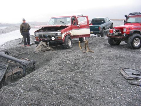 1994 Ford Bronco - A red and white Ford Bronco appears stuck in gravel, with a green pickup truck assisting. A red Jeep is also visible in the background.
