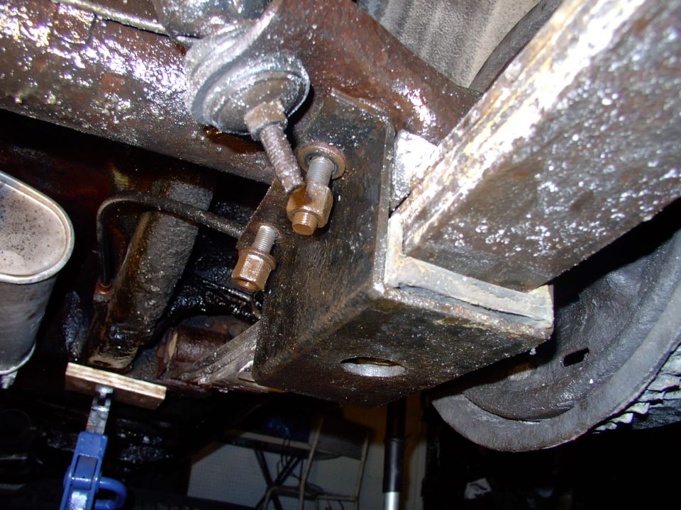 Undercarriage view of frame bracket with two bolts, grease-covered metal, brake line, and jack stand beneath.