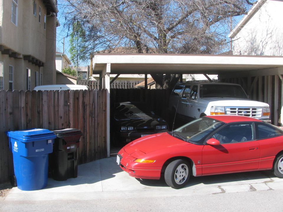 Red sports car parked in driveway, white SUV and black car under carport