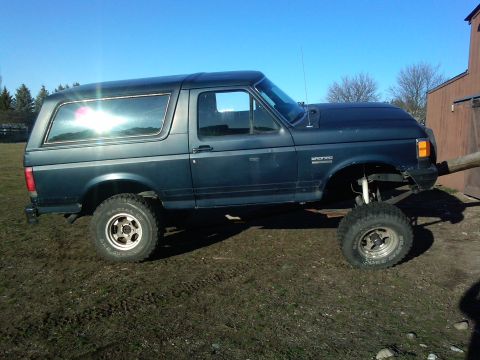 1988 Ford Bronco - Lifted Ford Bronco with aftermarket wheels and tires sits on muddy ground under a clear blue sky.