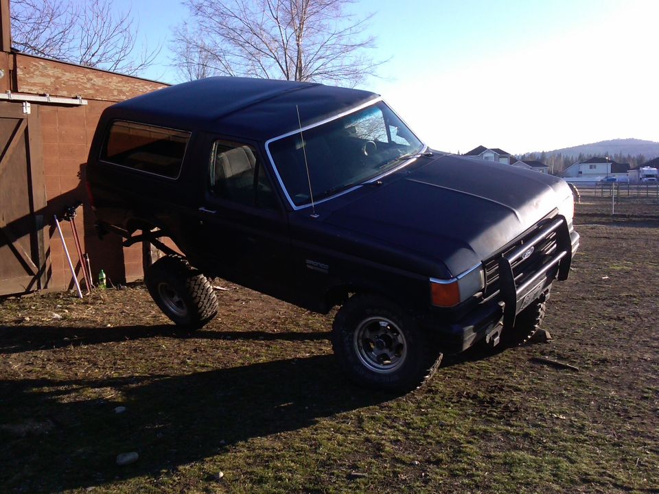 Dark blue Ford Bronco with one wheel lifted off the ground, parked on grass next to a wooden building