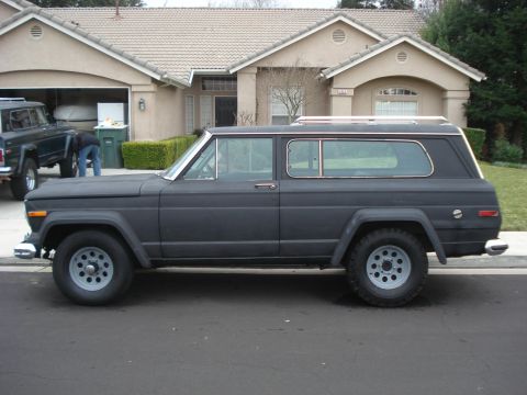 1977 Jeep Cherokee - A dark matte Jeep Wagoneer is parked on the street in front of a house, with another Jeep visible in the driveway.