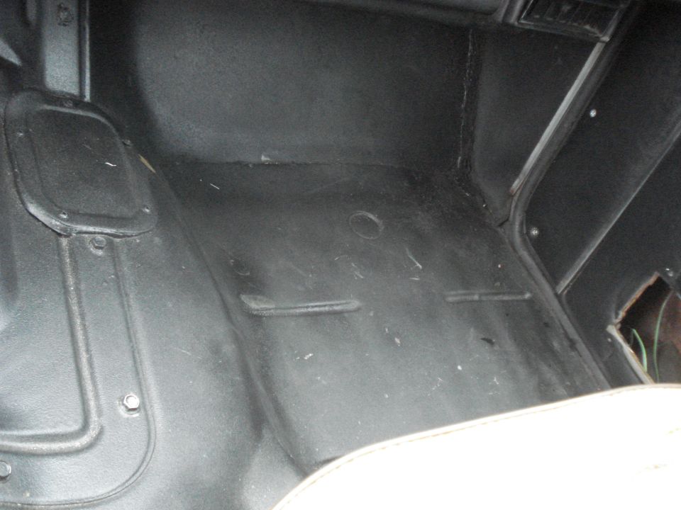 Black vehicle floor mat with raised ridges and a circular indentation. A white seat edge is visible at the bottom