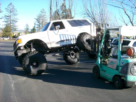 1989 Ford Bronco - A forklift is lifting a heavily modified white Ford Bronco, showing its lifted suspension and large tires.