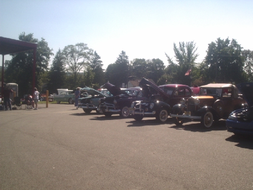 Several classic cars parked in a lot with hoods up, under a sunny sky