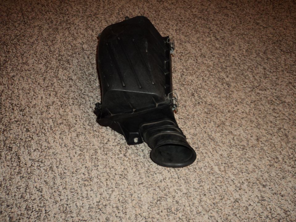 Black plastic air intake box with a flexible hose attached, lying on a carpeted surface