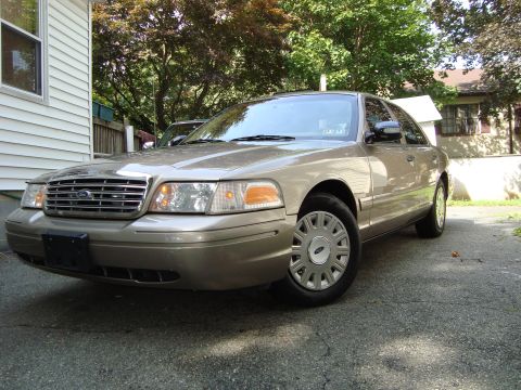 2004 Ford Crown Victoria - A tan 2004 Ford Crown Victoria is parked on a paved driveway in front of a residential building.