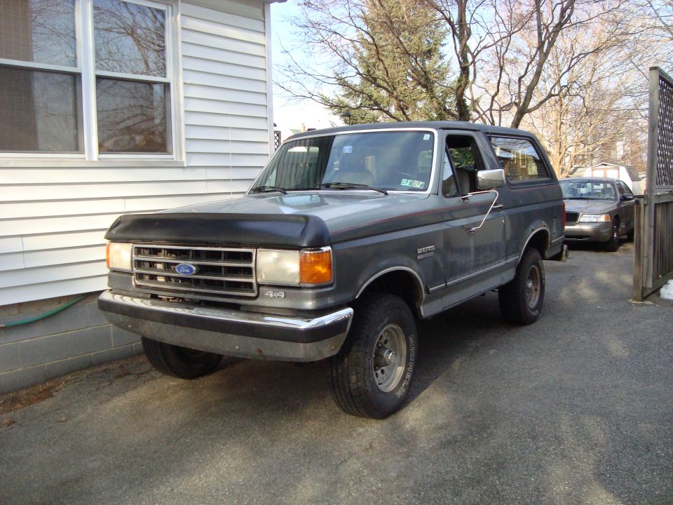 Dark gray Ford Bronco SUV with hood deflector and 4x4 badge parked in a driveway next to a white house