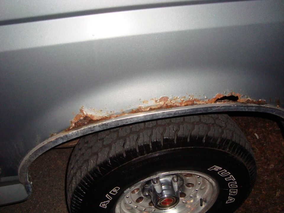 Close-up of a rusted-out rear wheel well on a vehicle. Tire is visible with 'FUTURE' written on it