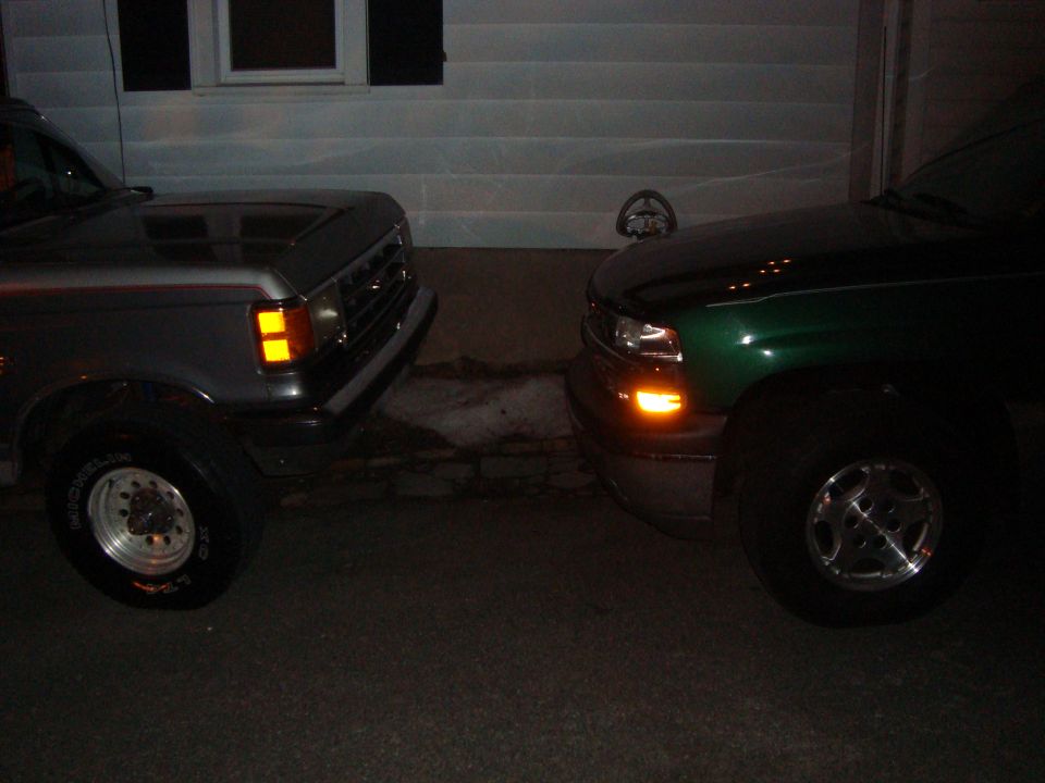 Gray truck on left, green truck on right, parked close together in front of a house