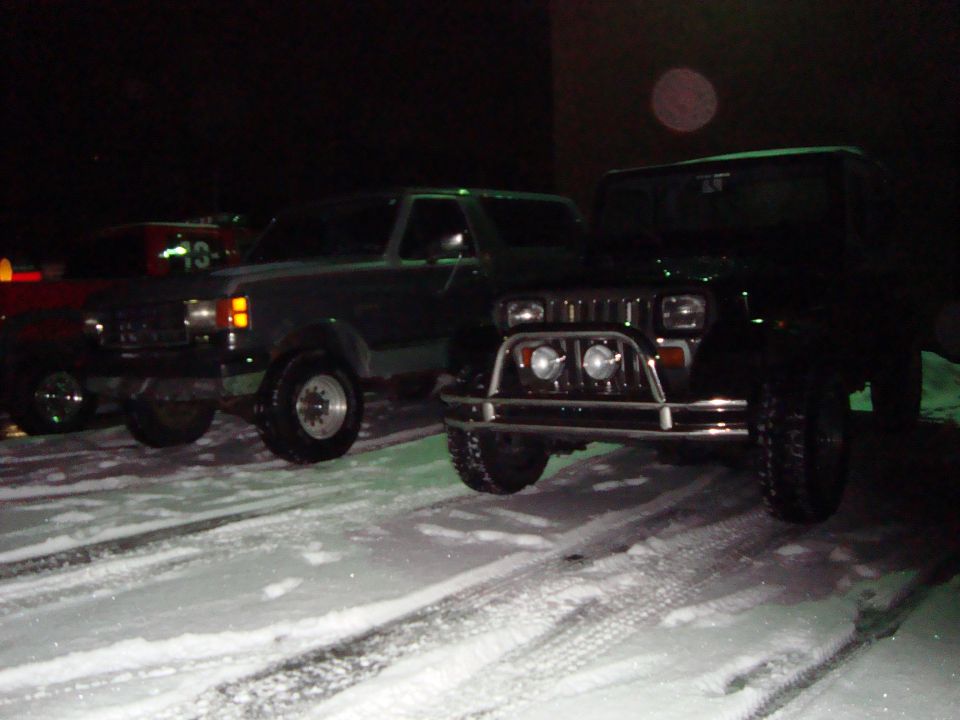 Dark green Ford Bronco and lifted Jeep with tube bumper parked in snow at night