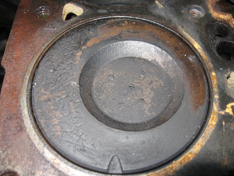 1981 Ford Bronco - Extreme close-up down a cylinder bore at a dished piston crown carrying carbon deposits and faint cast markings, with surface rust staining…
