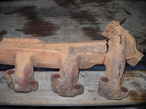 1993 Ford C-150 (Centurion Conversions) - A heavily rusted and cracked exhaust manifold is shown resting on wooden planks.