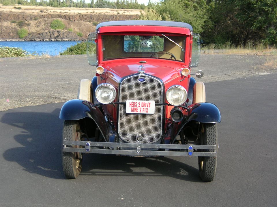 Red 1931 Ford Model A pickup truck front view with license plate reading "HERS 2 DRIVE MINE 2 FIX"