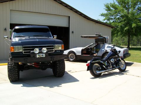 1990 Ford Bronco - A lifted dark blue truck sits in front of a garage with a DeLorean and a motorcycle parked nearby.