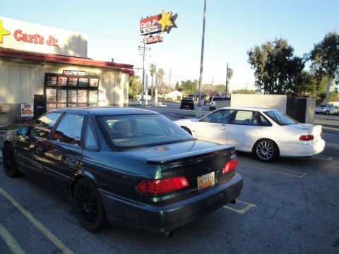 1993 Ford Taurus - A dark green 1993 Ford Taurus with aftermarket wheels is parked in a lot next to a white sedan.