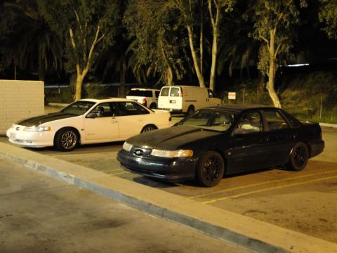 1993 Ford Taurus - Two sedans are parked side-by-side in a lot at night, with trees and vans visible behind them.