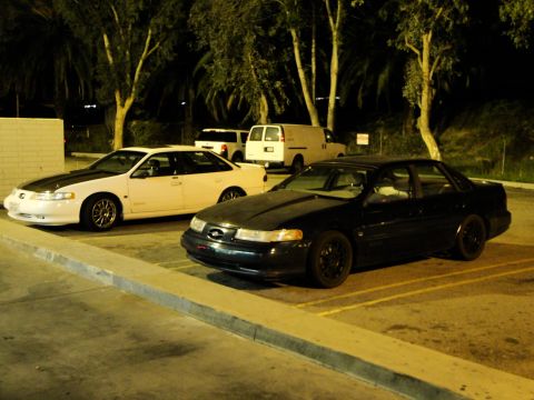 1993 Ford Taurus - Two sedans are parked in a lot at night, with vans visible in the background.