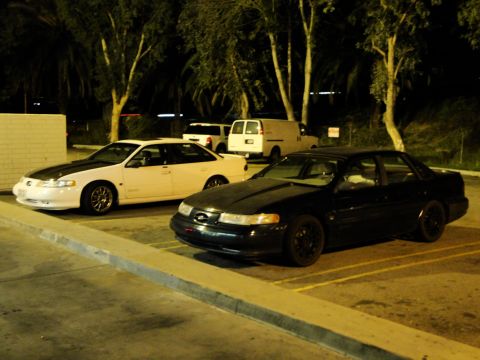1993 Ford Taurus - A black sedan with a modified hood and dark wheels is parked next to a white sedan at night.