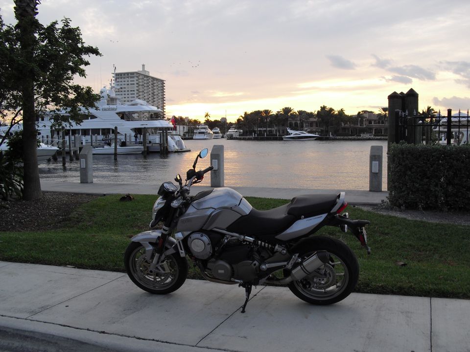 Silver naked motorcycle parked side-on by a marina at sunset with yachts behind.