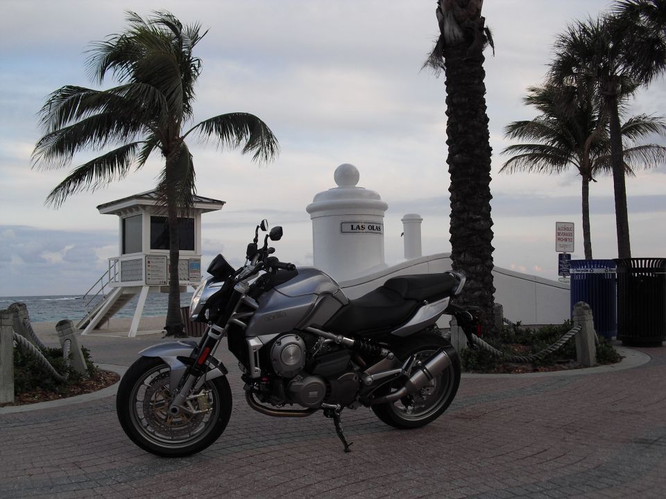 Silver and black Aprilia motorcycle parked on brick walkway near beach with palm trees