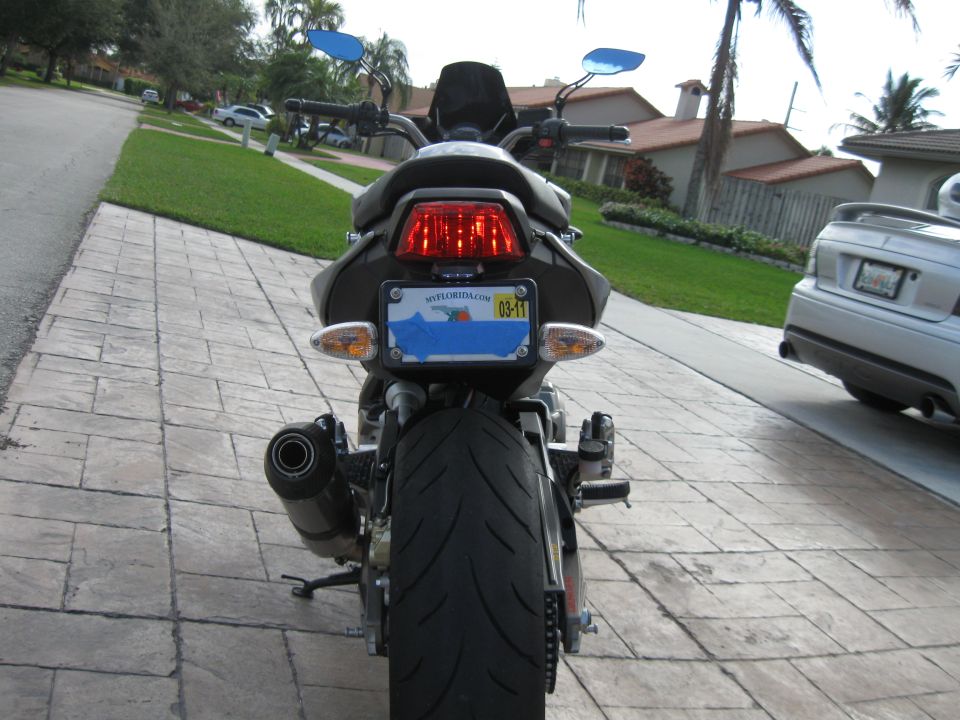 Rear view of a motorcycle on a driveway showing taillight, taped Florida plate, rear tire and exhaust