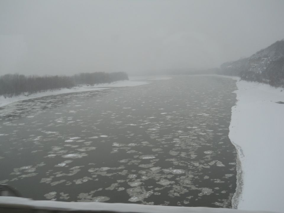 Frozen river with ice chunks floating, snow-covered banks, and a hazy sky