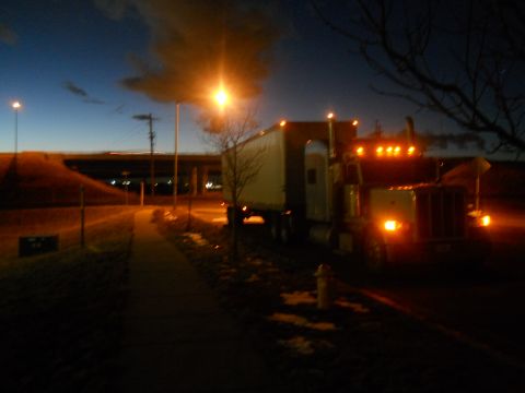 2009 Kenworth T800 - A yellow semi-truck with its lights on is parked on the side of the road during twilight.