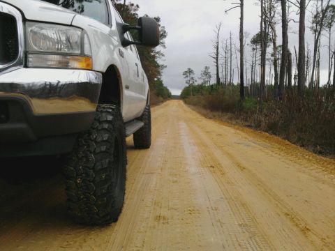 1995 Ford Bronco - A white pickup truck with off-road tires drives down a dusty dirt road through a wooded area.